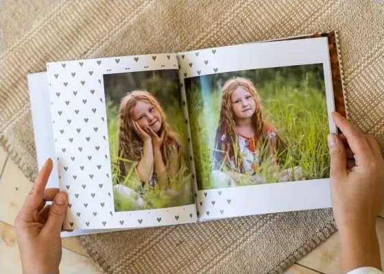 Руки держат открытую детскую фотокнигу с фото девочки в траве и узором из сердечек